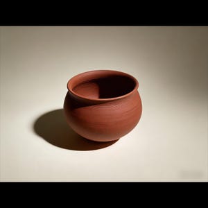 May include: A handmade, terracotta-colored ceramic bowl with a rounded body and a wide, open rim. The bowl has visible horizontal lines around its exterior, indicating the pottery process. The interior is dark, and the bowl casts a shadow on the neutral background.
