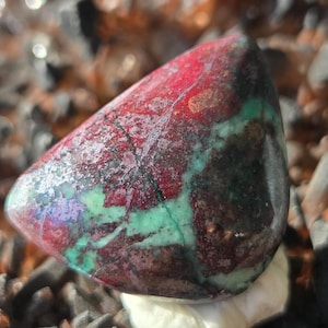 Puede incluir: Una piedra preciosa pulida con una mezcla de colores. La piedra presenta una mezcla de rojo intenso, verde oscuro y toques de morado. La superficie tiene un aspecto texturizado, con vetas y patrones visibles.