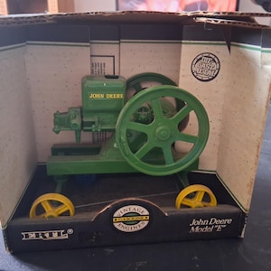 May include: A green John Deere Model "E" vintage gasoline engine model. The model has a large green flywheel, green engine components, and yellow wheels. The box reads "The Cast Metal" and "Vintage Gasoline Engines."