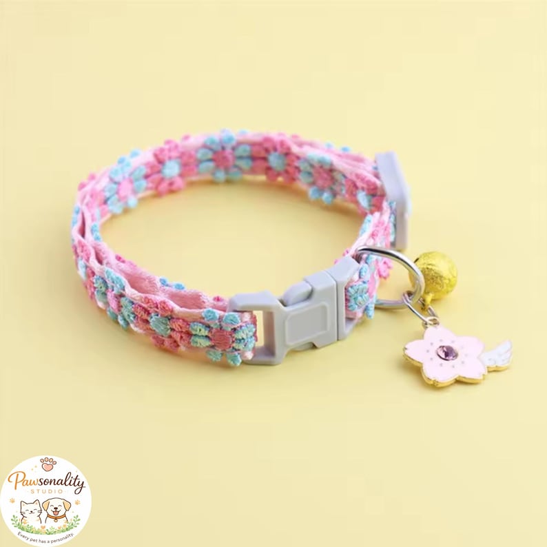 Pink and blue floral patterned pet collar with a gray buckle. The collar has a gold bell and a flower charm with a purple gem. The collar is designed for pets. The floral collar shown in a circular loop on a yellow background, highlighting the pink, blue, and white flower print.