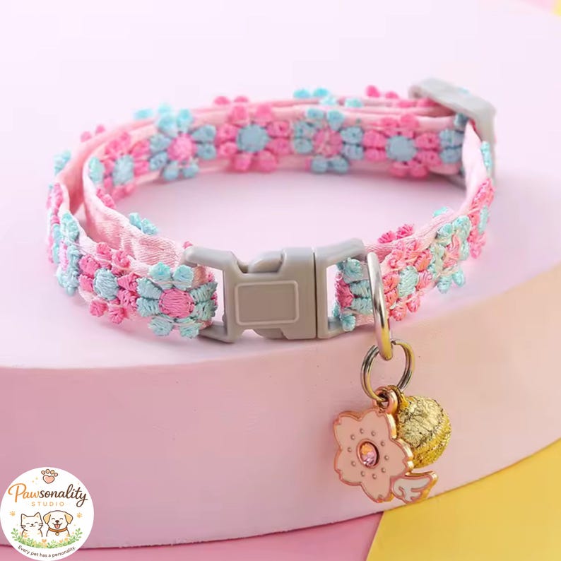 A close-up of the pink cat collar with a floral design in pink and blue. The collar has a gray plastic buckle and a gold-colored bell with a pink flower charm.