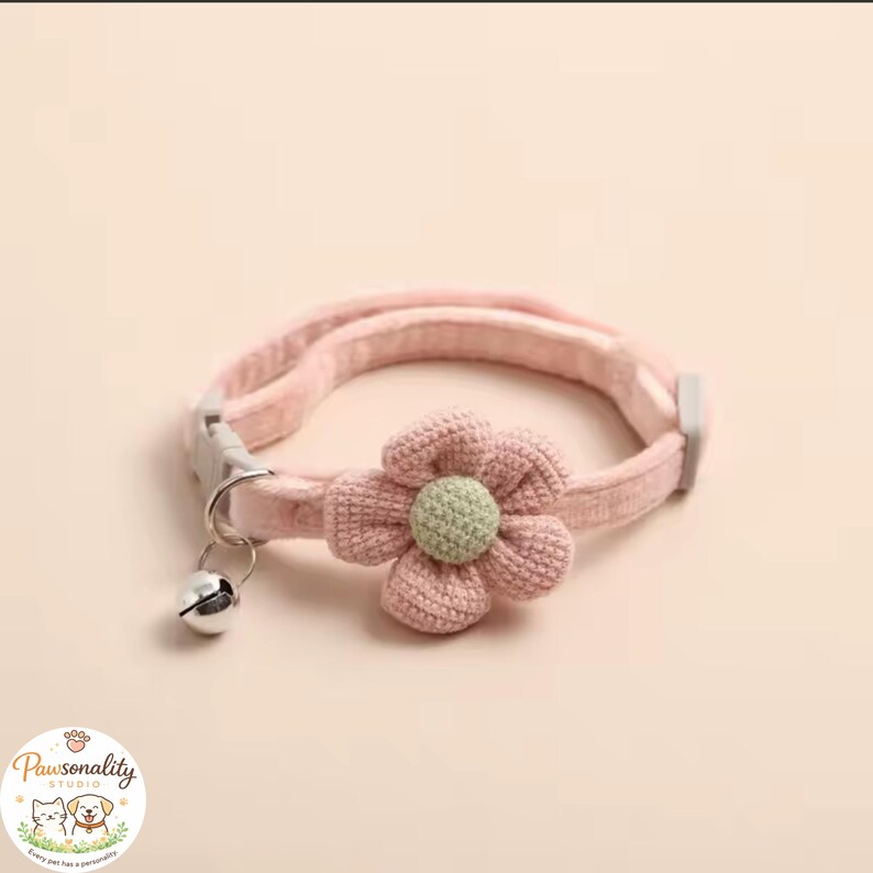 A close-up of the pink knit collar itself, laid flat against a cream background, featuring the pink flower with green center, a small silver bell, and a breakaway buckle