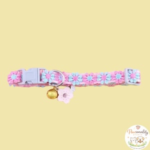 Pink and blue floral cat collar with a gold bell and a pink flower charm. The collar has a plastic buckle and a metal D-ring. The collar laid out flat horizontally, showing the adjustable strap, bell and flower bell attachment