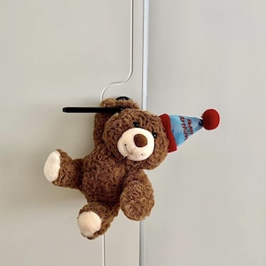 Puede incluir: Un oso de peluche marrón con un gorro de fiesta azul y rojo con las palabras "Happy Birthday". El oso tiene un hocico y patas blancas. El oso está colgado de una barra negra.