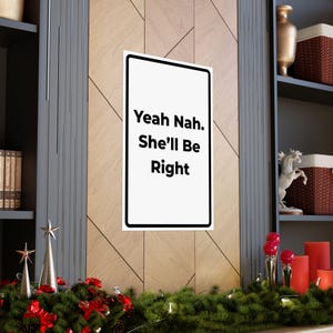 May include: A white rectangular sign with a black border displays the text "Yeah Nah. She'll Be Right" in a bold, sans-serif font. The sign is mounted on a wooden wall, with decorative shelves and holiday decor in the background.