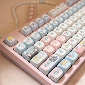 May include: A mechanical keyboard with a light pink frame and rounded keycaps in pastel shades of blue, pink, and white. The keys feature various anime-style character illustrations, symbols, and text including Tab, Caps, Shift, and Ctrl in a soft font.