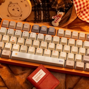 Puede incluir: Un teclado mecánico con marco naranja y teclas color hueso con letras naranjas. Las teclas incluyen iconos de sol y nube en la barra espaciadora y la tecla W. Las teclas de función F5, F6 y F7 son gris oscuro, con distribución QWERTY.