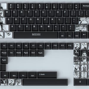 以下が含まれることがあります： サッカー選手の画像が特徴のカスタムキーキャップが付いた白黒キーボード。「MESSI」と書かれたスペースバー。キーボードは、数字キーとファンクションキーを備えた標準的なQWERTYレイアウトです。