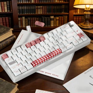 Puede incluir: Un teclado mecánico blanco con marco plateado y perilla giratoria. Las teclas muestran una mezcla de blanco, cuadros vichy rojos y blancos, y corazones rojos, con pequeñas ilustraciones de lazos rojos y el texto BackSpace.