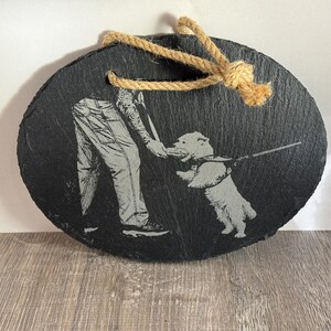 May include: Oval black slate ornament with a laser-etched design of a person playing with a dog. The dog is holding a stick. A natural rope is attached for hanging.