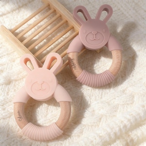 May include: Two pink bunny-shaped baby teethers. Each teether has a wooden ring with a textured silicone section and a bunny face. One teether has the name "Harry" and the other has "Glenn" engraved on the wooden ring.