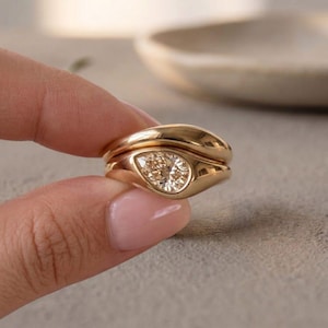 Op de afbeelding: Een gouden ring met een peervormige, gefacetteerde edelsteen in champagnekleur, gezet in een gladde, gepolijste metalen band. Het ontwerp bevat een tweede, effen gouden band die naadloos aansluit op de hoofdzetting voor een geheel.