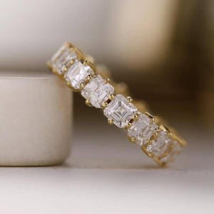 May include: A gold band ring featuring a continuous row of square-cut clear gemstones set in a prong setting. The ring displays a series of identical faceted stones arranged side by side, creating a uniform and reflective metallic and crystal design.
