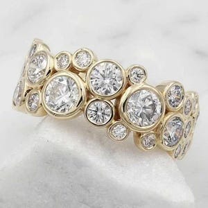Op de afbeelding: Een goudkleurige ring met een cluster van ronde, heldere edelstenen. De edelstenen variëren in grootte, waardoor een bubbelachtig, organisch ontwerp ontstaat. De ring ligt op een wit oppervlak, wat de glinstering van de stenen benadrukt.