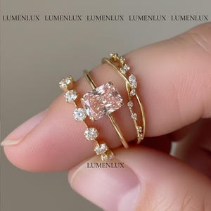 May include: Three gold rings are displayed, with a focus on the center ring featuring a large, rectangular, light pink gemstone. The other rings have smaller, clear gemstones. The word "LUMENLUX" is printed on the image.