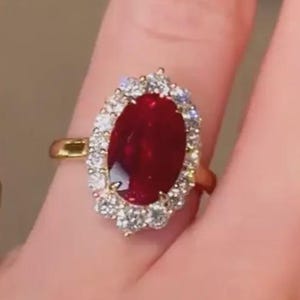 Puede incluir: Un anillo de oro con una gran piedra central ovalada roja, rodeada por un halo de pequeños diamantes. También es visible otro anillo de oro con una banda de diamantes. Las uñas están pintadas de rojo.