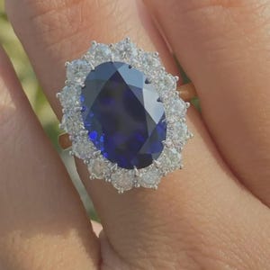 May include: An oval-cut, deep blue gemstone ring, surrounded by a halo of smaller, clear gemstones. The ring's setting appears to be gold, with the gemstones set in a silver or white metal. The ring is shown on a person's finger.