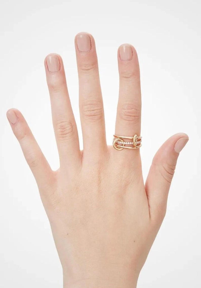 May include: A gold ring with an unusual design, comprising several interwoven bands. One band is set with small, sparkling stones. The ring is shown on a hand with neatly manicured nails, against a plain white backdrop.