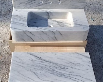 May include: A white marble bathroom sink with grey veining, featuring a rectangular basin and a backsplash with three tap holes. A matching marble shelf is positioned below the sink, all resting on a wooden support.
