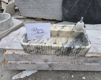 May include: A rectangular marble sink with a fluted exterior. The sink is white with grey veining and sits on a marble surface. A marble soap dispenser is on the right. A business card with the text "Rowan Marble" is in the centre.