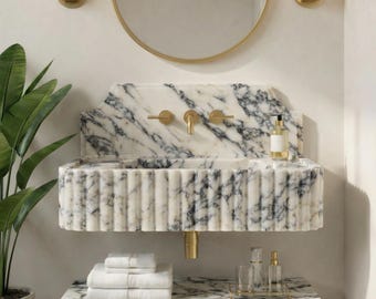 May include: A marble washbasin with a fluted design and gold taps. The basin is mounted on a marble shelf with white towels. A round mirror and gold sconces are mounted on the wall above the basin. A potted plant is on the left.
