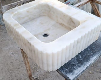 May include: A rectangular, white marble sink with a fluted exterior. The sink has a central drain hole and is set on a metal stand. The marble has subtle beige veining, adding to its natural aesthetic. The sink is a bathroom fixture.