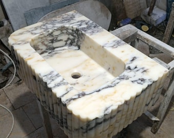 May include: A rectangular marble sink with a scalloped edge design. The sink features a cream and grey veined pattern, with a central drain hole. It is supported by a simple wooden stand, showcasing a natural stone aesthetic.