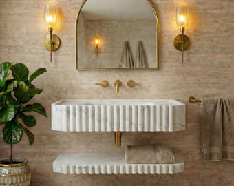 May include: A contemporary bathroom suite featuring a white marble sink and shelf with a fluted design. The set includes a gold-framed arched mirror, gold sconces, and a potted plant. A beige towel hangs on a gold towel rail.
