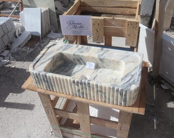 May include: A rectangular marble sink with a fluted exterior. The sink is white with grey veining and sits on a wooden stand. The sign in the background reads "Rowan Marble".
