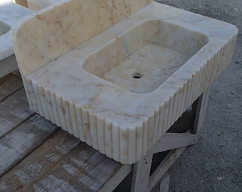May include: A rectangular marble sink with a backsplash. The sink is a light beige colour with darker veining. The front edge of the sink has a fluted design. The sink is sitting on a wooden stand.