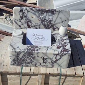 May include: A marble sink with a white and burgundy pattern. The sink has a rectangular basin, a back with two faucet holes, and a small soap dish. A white soap dispenser and a business card are on the side. The business card reads "Rowan Marble".
