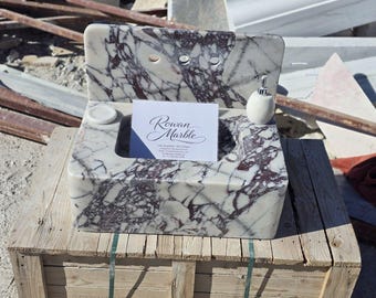 May include: A marble sink with a white and burgundy pattern. The sink has a rectangular basin, a back with two faucet holes, and a small soap dish. A white soap dispenser and a business card are on the side. The business card reads "Rowan Marble".