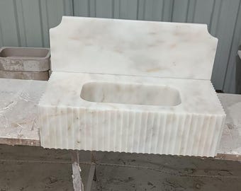 May include: A white marble bathroom sink with a rectangular basin and a scalloped edge. The sink has a back splash and is supported by a metal frame. The marble has a natural veining pattern.