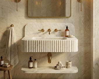 May include: A contemporary bathroom sink crafted from white marble with a fluted pattern. It features gold-toned taps and a waste. A marble shelf below holds toiletries and rolled towels. The walls are marble, and the lighting fixtures are gold.