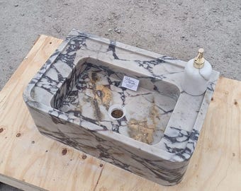 May include: A rectangular marble sink with a grey, black, and gold veined pattern. A white soap dispenser with a gold pump sits on the right side of the sink. The sink is on a wooden pallet.