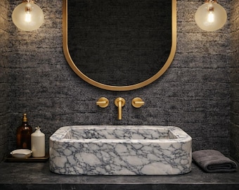 May include: A contemporary bathroom featuring a marble sink, gold taps, and an oval mirror. Two spherical wall lights are positioned either side of the mirror. The sink is rectangular with rounded edges. The wall is dark grey stone.