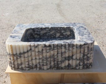 May include: A rectangular marble sink with a carved, fluted exterior. The sink basin is a deep rectangle. The marble has a white base with grey veining. The sink is displayed on a light-coloured wooden surface.