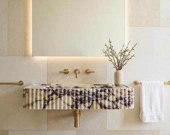 May include: A contemporary bathroom featuring a marble sink with a ribbed design and purple veining. The sink has gold fixtures, including a tap and drain. A rectangular mirror with a light frame hangs above. A white towel hangs on a gold rail. A vase with branches sits on the sink.