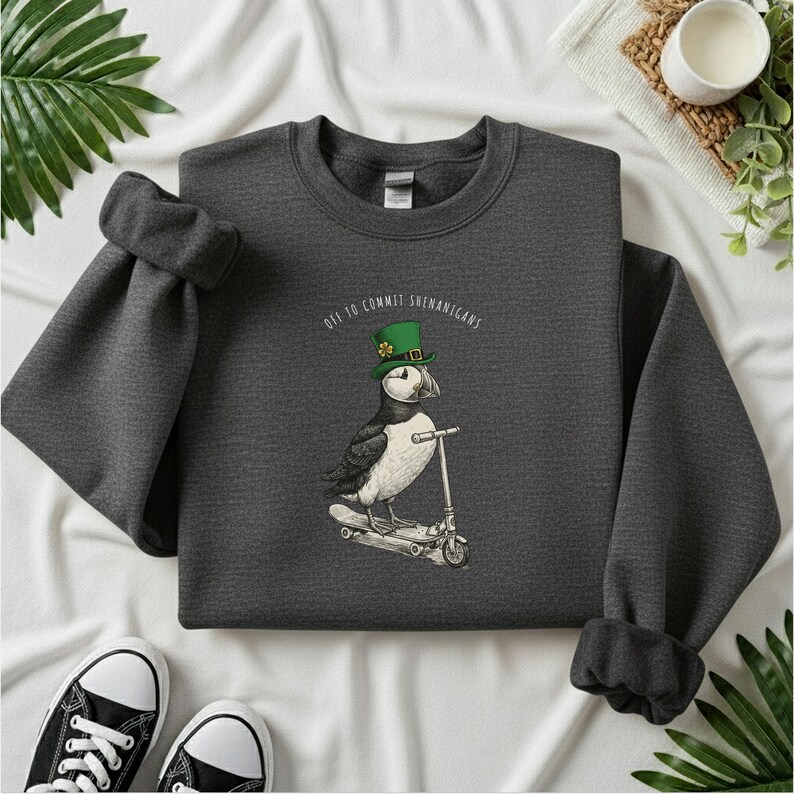 Może przedstawiać: Ciemnoszara bluza z maskonurem w zielonym kapeluszu jadącym na hulajnodze. Nad maskonurem widnieje napis "Off to Commit Shenanigans". Czarne i białe trampki widoczne na dole zdjęcia.