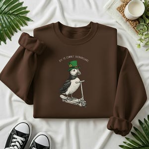 Może przedstawiać: Brązowa bluza z nadrukiem maskonura w zielonym kapeluszu na skuterze. Nadruk zawiera napis "Off to Commit Shenanigans". Czarne i białe trampki u dołu.