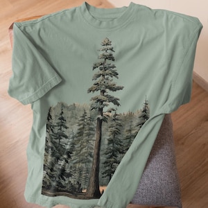 May include: Sage green t-shirt featuring a detailed illustration of a tall pine tree in a forest. The artwork showcases a realistic depiction of the tree and surrounding trees, with a focus on the natural environment. The shirt is made of soft material.