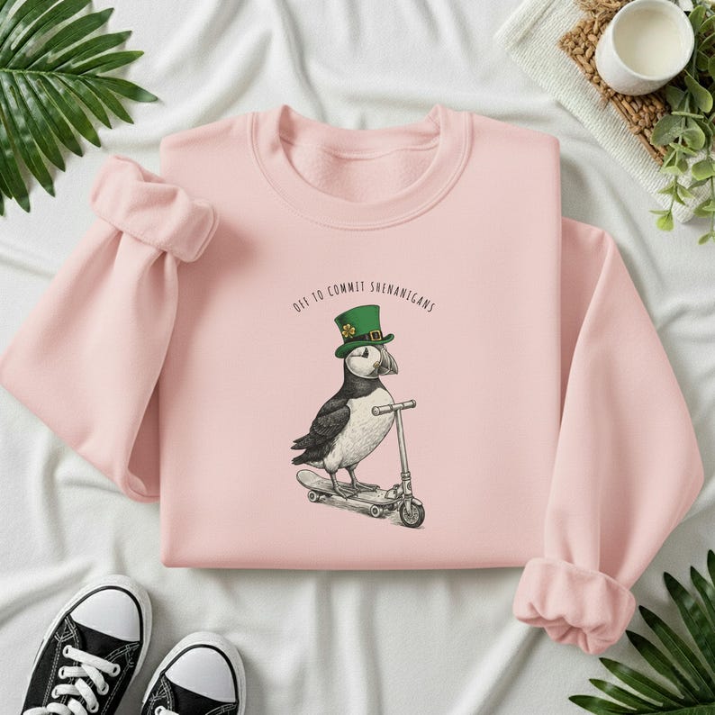 Może przedstawiać: Jasnor&oacute;żowa bluza z napisem "Off to Commit Shenanigans". Grafika przedstawia maskonura w zielonym kapeluszu na skuterze. Czarne i białe trampki widoczne na dole zdjęcia.