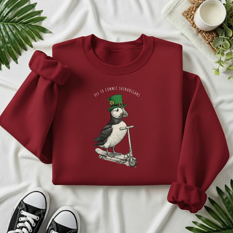Może przedstawiać: Bordowa bluza z nadrukiem maskonura w zielonym kapeluszu krasnoludka na hulajnodze. Tekst "Off to Commit Shenanigans" jest wygięty nad maskonurem. Czarne i białe trampki widoczne na dole.
