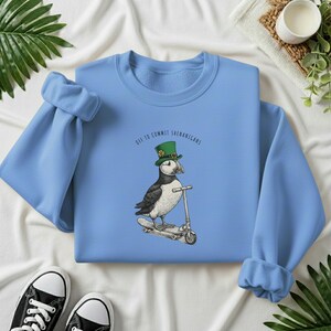 Może przedstawiać: Jasnoniebieska bluza z maskonurem w zielonym kapeluszu na hulajnodze. Nad maskonurem widnieje napis "Off to Commit Shenanigans". Czarne i białe trampki widoczne na dole zdjęcia.