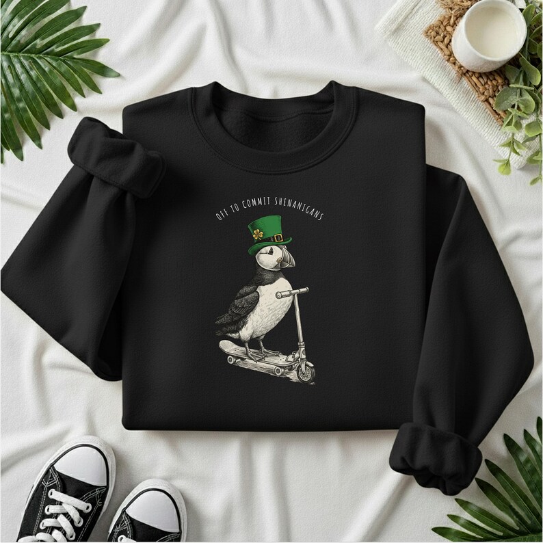 Może przedstawiać: Czarna bluza z maskonurem w zielonym kapeluszu jadącym na hulajnodze. Tekst "Off to Commit Shenanigans" znajduje się nad maskonurem. Czarno-białe trampki znajdują się na dole obrazu.