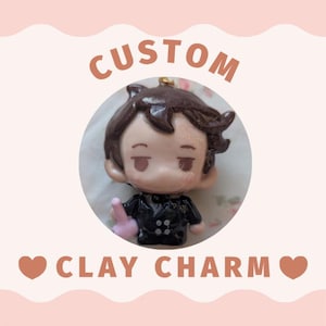 May include: A custom clay charm featuring a small, stylized figure with dark brown hair, wearing a black suit with white buttons. The figure has a neutral expression and holds a small pink object. The text CUSTOM CLAY CHARM is visible.