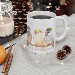 May include: White ceramic mug featuring a charming illustration of three colorful birds perched on a branch. The birds have pink beaks and are adorned with a bow and a hat. The mug is filled with a dark liquid.
