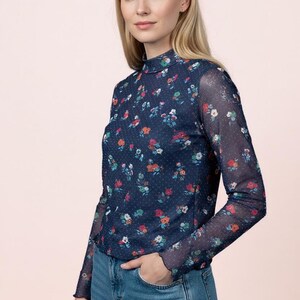 Puede incluir: Una blusa de manga larga con cuello alto confeccionada en tejido de malla azul marino transparente, con un pequeño patrón de lunares blancos y un estampado floral colorido con flores rojas, naranjas y verdes. La prenda tiene un corte ajustado.