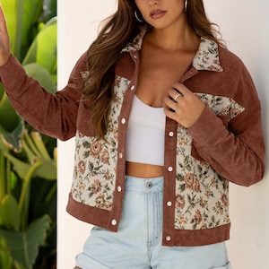 May include: A brown corduroy jacket with floral print panels and a white top. The jacket has a button-up front and a collared neckline. The floral print features pink, beige, and green flowers on a cream background. Light blue denim shorts complete the outfit.