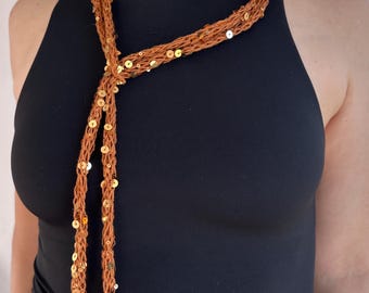 May include: A long, thin, rust-colored scarf made of a loose, open-knit yarn material. The scarf is embellished with numerous small, reflective gold-toned sequins scattered throughout the length of the fabric, creating a shimmering effect.